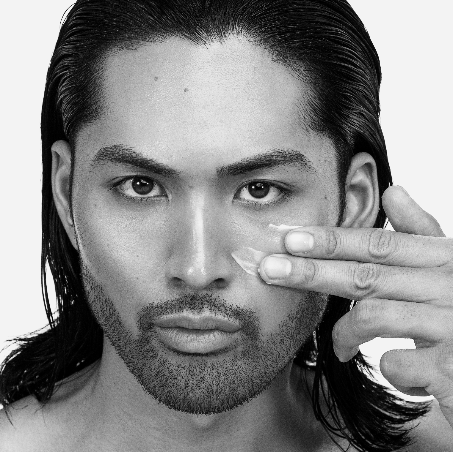 Black and white portrait of a person with long hair and a beard, applying skincare to their face.